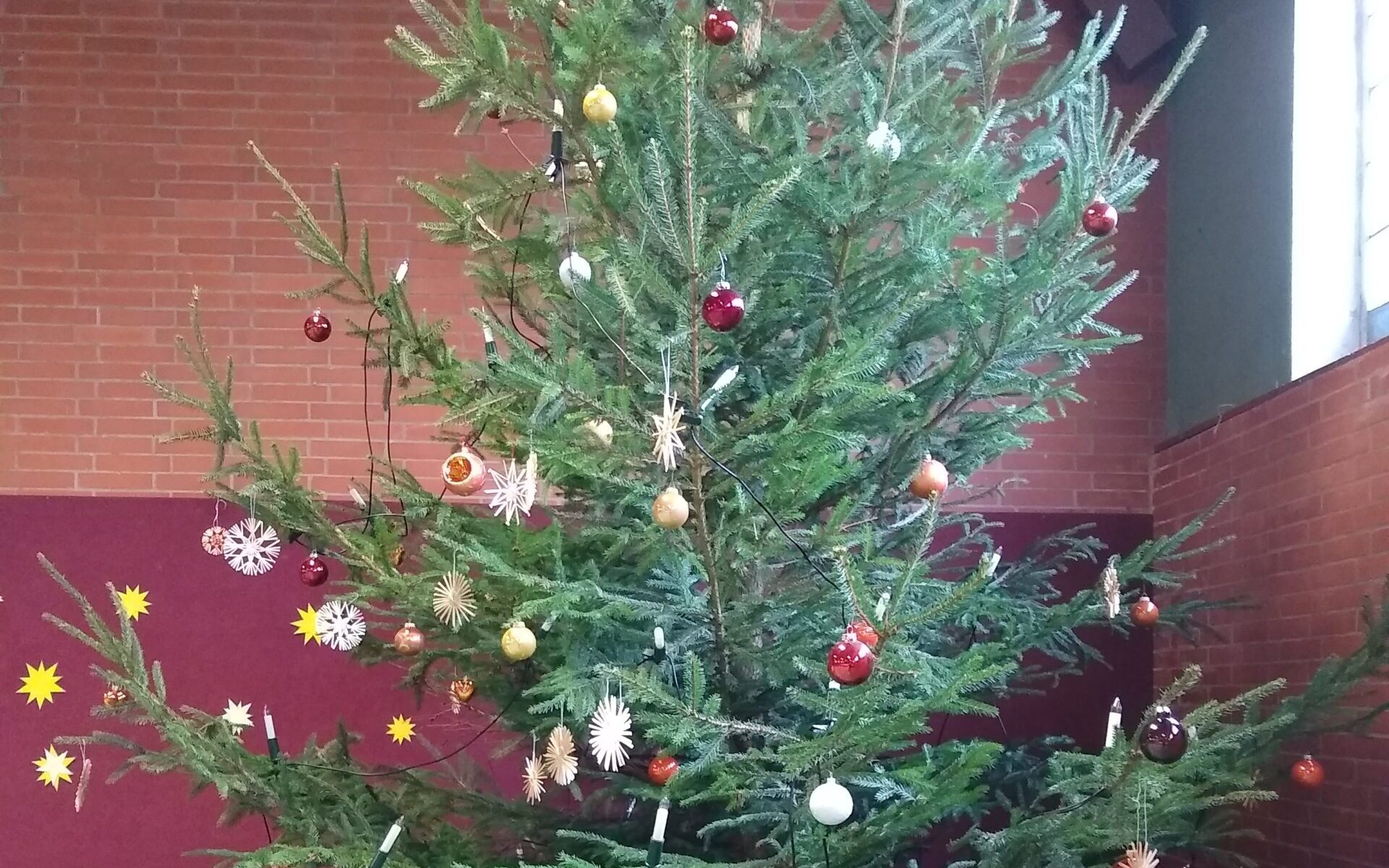 Ein geschmückter Weihnachtsbaum in einer Gemeindehalle in Sehlde, aufgenommen im Jahr 2019. Der Baum ist mit Kugeln, Strohsternen und Lichterketten dekoriert, unter dem Baum liegen verpackte Geschenke.